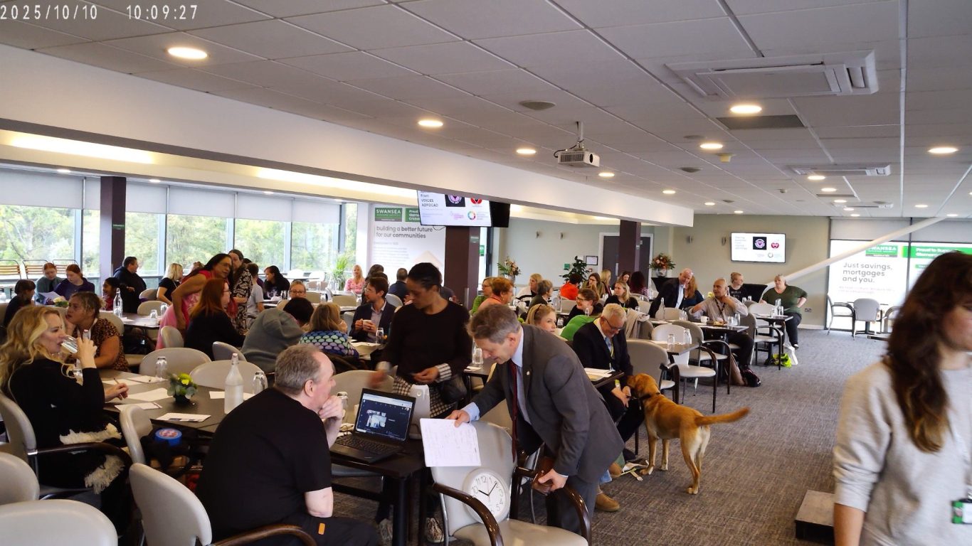 A busy indoor event with people working at tables and a dog among them.